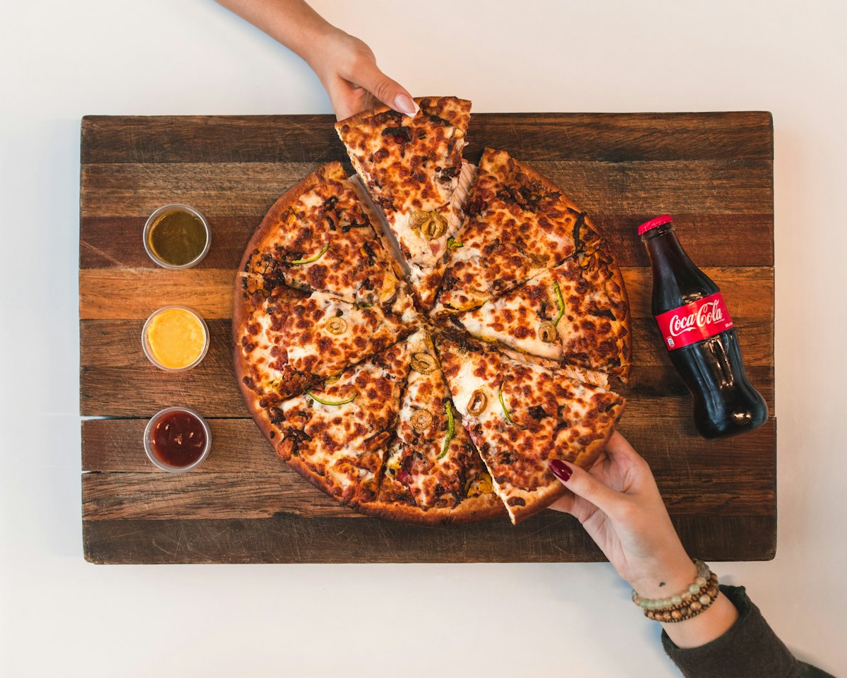 Pizza fatiada sobre tábua de madeira, com molhos e refrigerante, sendo dividida entre amigos